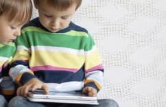 Two children in colorful striped sweaters using a tablet, sitting on a white textured surface.