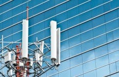 Cell tower with white antennas against a blue glass building under a clear sky.