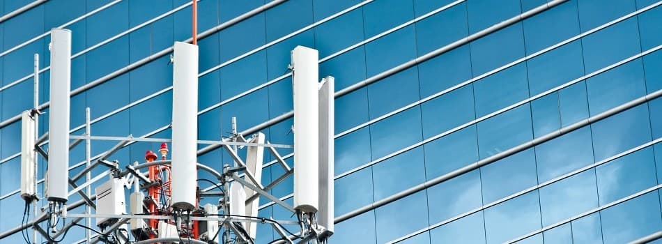 Cell tower with white antennas against a blue glass building under a clear sky.