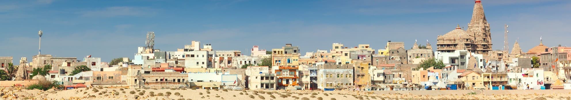 Panoramic view of a desert town with colorful buildings, a cell tower, and a large temple with spires under a clear blue sky, set against a sandy foreground.